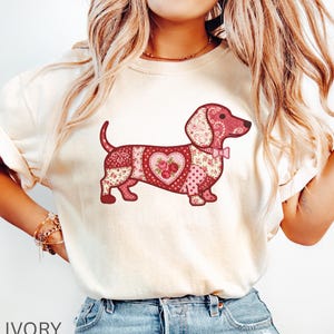 Puede incluir: Camiseta de color marfil con un diseño de perro salchicha en patchwork. El perro está compuesto por varios patrones florales rosas y rojos, con un corazón en el centro y una pequeña pajarita. La palabra "IVORY" está impresa en la parte inferior.