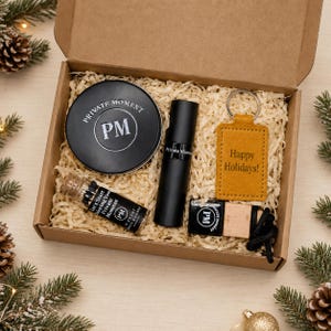 May include: A cardboard gift box containing a black circular tin labeled Private Moment PM, a black cylindrical bottle, a small glass vial with a cork stopper, a mustard yellow rectangular leather keychain with Happy Holidays! and a black diffuser.
