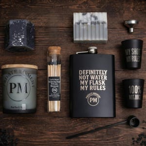 May include: A collection of lifestyle items arranged on a wooden surface. Includes a black flask with text, a candle, matches, soap, shot glasses, and a scoop. The flask reads "DEFINITELY NOT WATER MY FLASK MY RULES."
