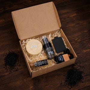May include: A gift set in a cardboard box with wood shavings. The set includes a wooden jar, a black spray bottle, a small glass bottle, a black keychain, and a wooden car air freshener. All items have the initials "PM".