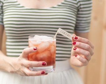 May include: A glass of pink iced drink with ice cubes in a clear glass. A wooden stir stick with a paper flag that says "CHEERS" is in the drink.