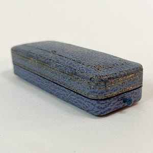 May include: A small, rectangular jewelry box in a muted blue hue with a textured surface. The box is trimmed with gold-colored accents and features a small, round blue clasp. The box is closed.
