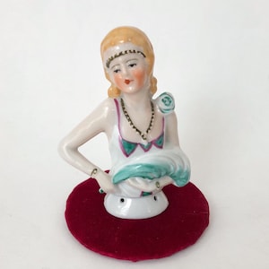 May include: A vintage porcelain figurine of a woman's bust, featuring blonde hair, a white dress with green and pink accents, and a pearl necklace. The figurine is mounted on a round, red velvet base. The bust is a decorative item.