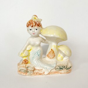 May include: A ceramic figurine of a mermaid with a yellow and brown mushroom. The mermaid has a blue tail and is holding a smaller mushroom. The figurine is painted in shades of yellow, brown, blue, and white.