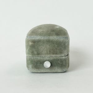 May include: A sage green velvet ring box with a rounded top and a rectangular base. The box features a single, small, circular white button closure centered on the front panel, providing a secure mechanism for keeping the lid closed tightly.