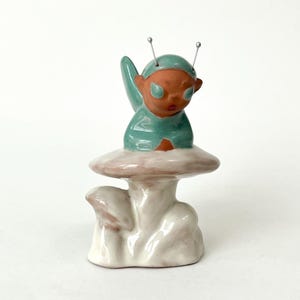 May include: A whimsical ceramic figurine of an alien-like creature atop a mushroom. The figure has a teal outfit and a terracotta-colored face with large eyes. The mushroom has a white stem and a pinkish-beige cap. Two silver antennae extend from the figure's head.