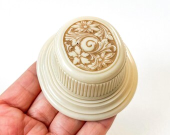 May include: A small, oval-shaped, cream-colored trinket box with a raised, floral design in a brown color. The box has a smooth, glossy finish and a decorative, ridged base.