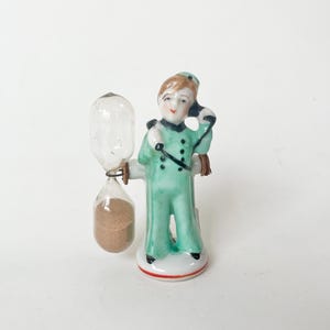 May include: A porcelain figurine of a person wearing a light green uniform with dark accents, holding a black telephone receiver to their ear. Attached to the side of the figure is a glass hourglass containing brown sand, set on a round base.