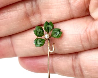 May include: A gold-toned hat pin featuring a four-leaf clover design. The clover has green enamel leaves with white veining and a small pearl in the center. The pin is held in a hand.