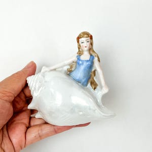 May include: A porcelain figurine of a woman with brown hair and a blue dress sitting on a white seashell.