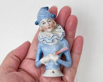 May include: A porcelain figurine of a jester with a light blue hat and outfit, a white ruffled collar, and a pink and brown object in its hands. The figurine has a painted face and is held in a hand.