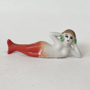 May include: A small porcelain mermaid figurine. The mermaid has a red tail, a white torso, and brown hair. Her arms are raised, and she has green accents on her ears. The figurine is lying down.