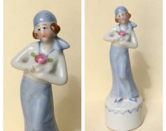 May include: A porcelain figurine of a person wearing a blue headscarf and a long blue skirt. The figure has brown hair, a white top with a pink rose accent, and stands on a cylindrical white base with a blue zigzag pattern around the top edge.