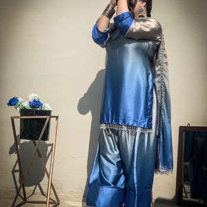 Puede incluir: Un conjunto de salwar kameez de satén azul y plateado. El atuendo presenta una túnica larga con efecto degradado, pantalones de pernera ancha y una bufanda a juego. Adornos plateados adornan el dobladillo de la túnica y la bufanda. El conjunto se combina con bailarinas plateadas.
