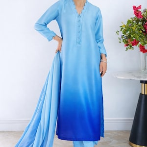 May include: A light blue two-piece outfit with a long-sleeve top and matching pants. The top features a V-neckline with delicate trim and a gradient effect, transitioning to a darker blue at the hem. The outfit is paired with light blue pants and silver heels.