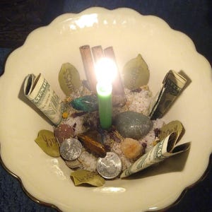 Puede incluir: Un cuenco crema con borde festoneado con una vela verde encendida en el centro. Billetes enrollados, monedas, piedras y palitos de canela rodean la vela. El cuenco contiene arena y peque&ntilde;os objetos con forma de hoja con texto.
