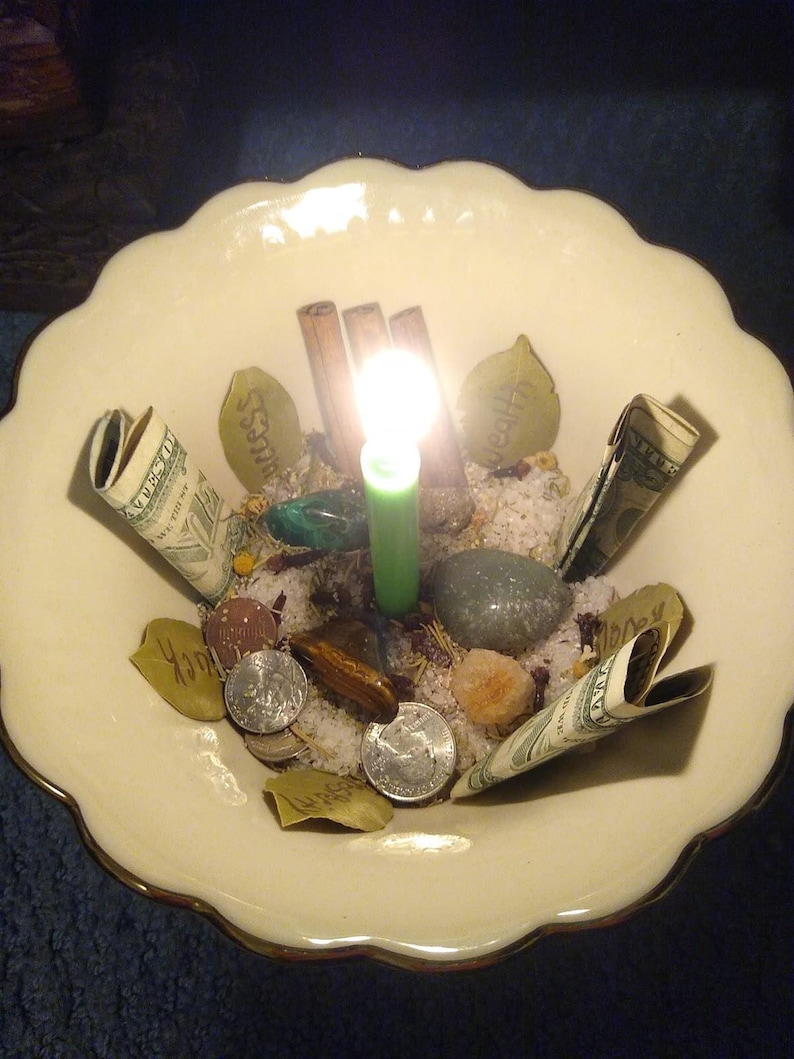 Puede incluir: Un cuenco crema con borde festoneado con una vela verde encendida en el centro. Billetes enrollados, monedas, piedras y palitos de canela rodean la vela. El cuenco contiene arena y peque&ntilde;os objetos con forma de hoja con texto.