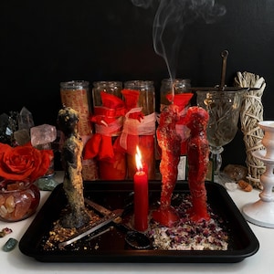 May include: A black tray holds a black statue, red candles, and a pair of red figure candles. A lit red candle and a pair of red figure candles are in the center. Dried herbs and rose petals are scattered around the candles. Smoke rises from one of the candles.