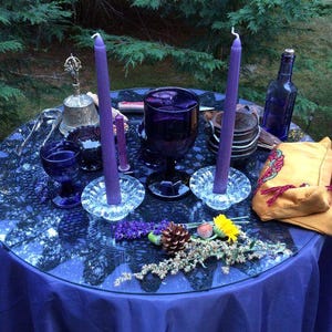 May include: A table set with various items, including purple candles, goblets, and a bell. The table is covered with a blue cloth and decorated with flowers, a pinecone, and a small yellow pouch. A blue glass bottle is also present.