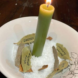 Puede incluir: Una vela verde encendida en un cuenco blanco con sal, hojas de laurel y peque&ntilde;os trozos de madera. La vela tiene una parte superior amarilla y una llama. El texto en las hojas dice "defy gravity" y "success in my interview".