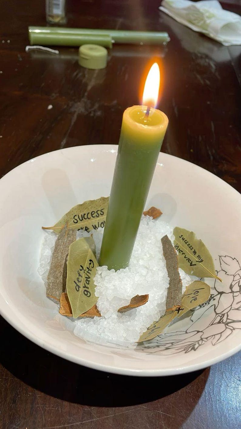 Puede incluir: Una vela verde encendida en un cuenco blanco con sal, hojas de laurel y peque&ntilde;os trozos de madera. La vela tiene una parte superior amarilla y una llama. El texto en las hojas dice "defy gravity" y "success in my interview".