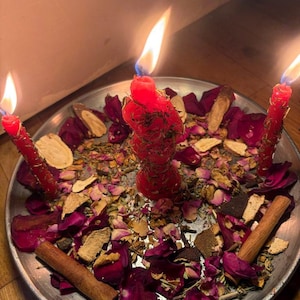 May include: A silver plate holds three burning red candles, surrounded by dried rose petals, herbs, and cinnamon sticks. The candles have long, flickering flames. The arrangement suggests a ritual or spiritual practice.
