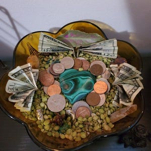 Puede incluir: Un cuenco decorativo de vidrio con borde dorado lleno de guisantes verdes, monedas y billetes doblados. Una gran piedra de malaquita verde y negra se encuentra en el centro. Varios cristales y peque&ntilde;os objetos est&aacute;n dispuestos alrededor del cuenco.