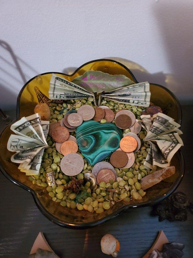 Puede incluir: Un cuenco decorativo de vidrio con borde dorado lleno de guisantes verdes, monedas y billetes doblados. Una gran piedra de malaquita verde y negra se encuentra en el centro. Varios cristales y peque&ntilde;os objetos est&aacute;n dispuestos alrededor del cuenco.