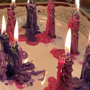 May include: Lit candles in shades of purple and pink, with melted wax pooling on a white plate. The candles have a twisted design and are adorned with silver glitter. The flames are bright and the scene is softly lit.