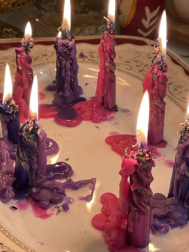 May include: Lit candles in shades of purple and pink, with melted wax pooling on a white plate. The candles have a twisted design and are adorned with silver glitter. The flames are bright and the scene is softly lit.