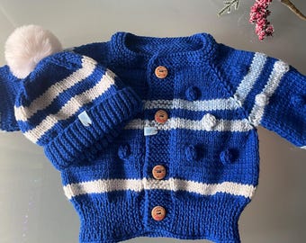 May include: A knitted baby set in blue and white, featuring a button-down cardigan and a matching beanie with a pom-pom. The cardigan has wooden buttons and textured details. The set is designed with striped patterns.