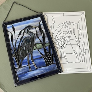 May include: Stained glass panel featuring a heron in a marsh scene with blue, green, and black colors. The artwork is framed in a dark blue border and includes a hanging chain. A line drawing of the heron is next to the panel.