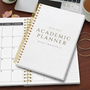 May include: A white academic planner with a gold spiral binding, open to a weekly calendar page. The cover reads "2026-2027 Academic Planner Weekly & Monthly" in gold lettering. The planner is on a wooden surface.
