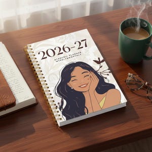 Op de afbeelding: Een spiraalgebonden academische planner voor 2026-27 met een illustratie van een lachende vrouw op de omslag. De omslag bevat ook de tekst "Academic Planner Weekly & Monthly". Een koffiemok en een bril staan in de buurt.