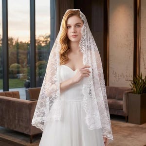 May include: A white bridal veil with intricate lace detailing. The veil is draped over a white wedding dress. The image is taken indoors with natural light, showcasing the delicate texture and design of the veil.