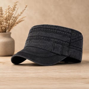 May include: A charcoal black military-style cadet cap featuring a flat top, a curved brim, and multiple rows of horizontal stitching across the crown. The fabric has a washed, faded appearance with a single metal eyelet visible on the side panel.