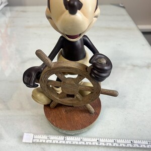 May include: A Mickey Mouse figurine holding a wooden ship's wheel. The figure has a tan face and ears, a black body, and yellow pants. The base is brown with the words "Disney World" printed on it.