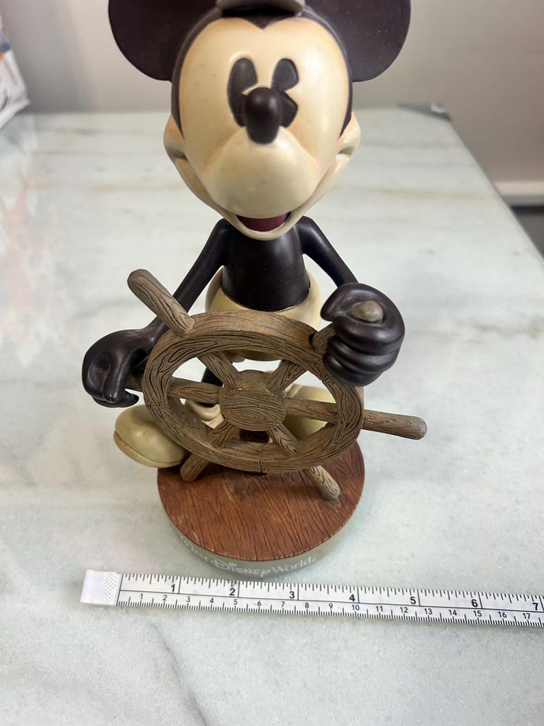 May include: A Mickey Mouse figurine holding a wooden ship's wheel. The figure has a tan face and ears, a black body, and yellow pants. The base is brown with the words "Disney World" printed on it.