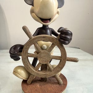 May include: A Mickey Mouse figurine in a sailor outfit, holding a wooden ship's wheel. The figurine has a cream-colored face and body, with black ears and a dark brown outfit. The base reads "Walt Disney World."