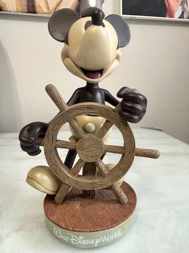 May include: A Mickey Mouse figurine in a sailor outfit, holding a wooden ship's wheel. The figurine has a cream-colored face and body, with black ears and a dark brown outfit. The base reads "Walt Disney World."