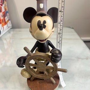 May include: A Mickey Mouse figurine in a sailor outfit, holding a wooden ship's wheel. The figure has a black body, tan face, and a gray hat. The base is brown with the text "Disney World".