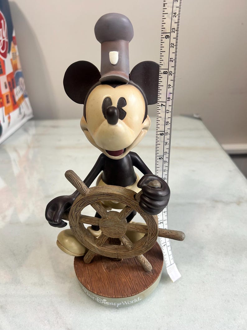 May include: A Mickey Mouse figurine in a sailor outfit, holding a wooden ship's wheel. The figure has a black body, tan face, and a gray hat. The base is brown with the text "Disney World".
