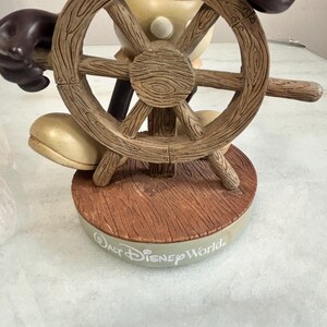 May include: A figurine of a cartoon character holding a wooden ship's wheel. The figurine is brown, beige, and black, with the words "Walt Disney World" on the base. The wheel has a detailed wood grain texture.