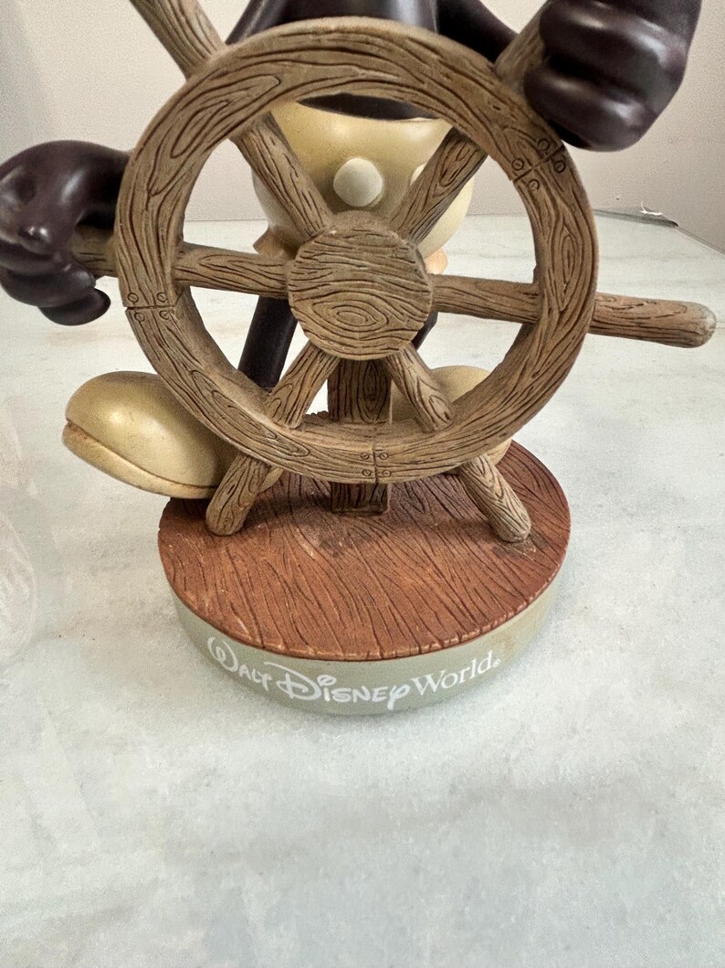 May include: A figurine of a cartoon character holding a wooden ship's wheel. The figurine is brown, beige, and black, with the words "Walt Disney World" on the base. The wheel has a detailed wood grain texture.