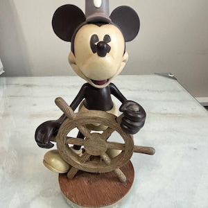 May include: A Mickey Mouse figurine in a sailor's outfit, holding a wooden ship's wheel. The figure has a tan face, black ears, and a dark gray hat. The base is brown with "Walt Disney World" printed on it.
