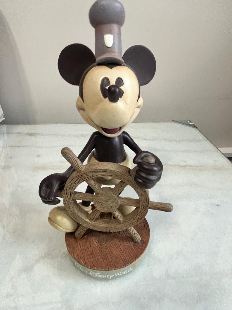 May include: A Mickey Mouse figurine in a sailor's outfit, holding a wooden ship's wheel. The figure has a tan face, black ears, and a dark gray hat. The base is brown with "Walt Disney World" printed on it.