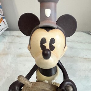 May include: A Mickey Mouse figurine in a chef's hat, holding a ship's wheel. The figure has a cream-colored face and body, with black ears, arms, and a nose. The chef's hat is gray, and the ship's wheel is a light brown color.