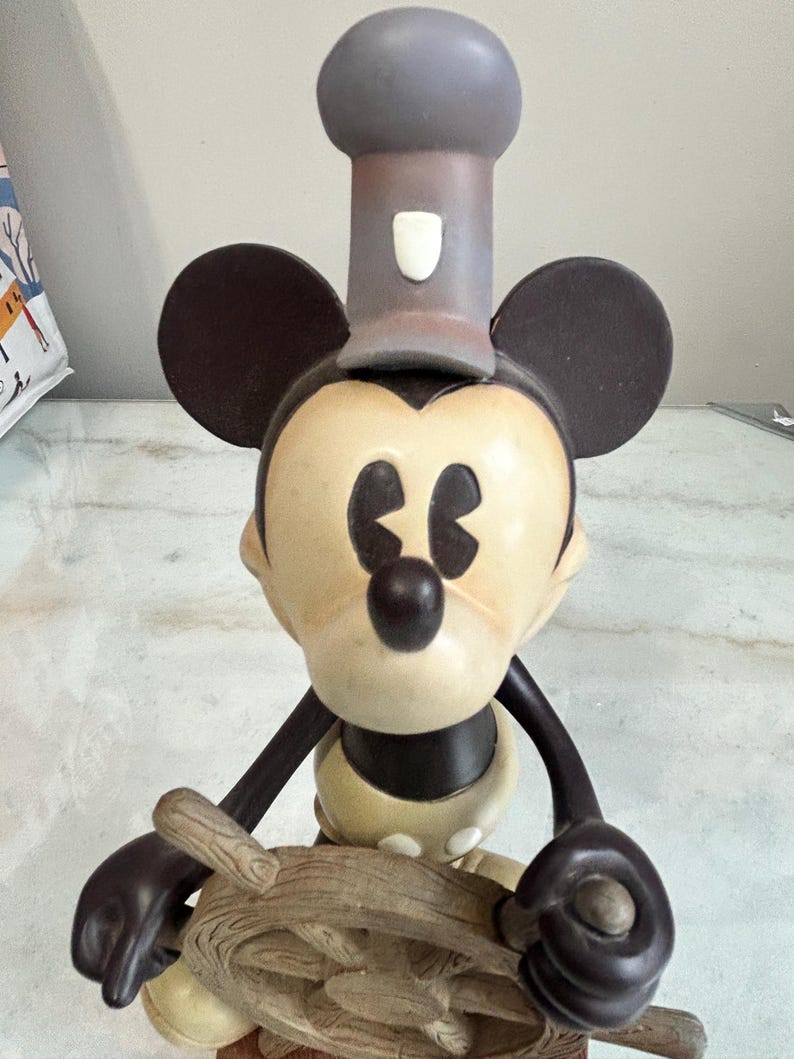 May include: A Mickey Mouse figurine in a chef's hat, holding a ship's wheel. The figure has a cream-colored face and body, with black ears, arms, and a nose. The chef's hat is gray, and the ship's wheel is a light brown color.