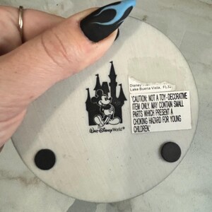 May include: A round, off-white Disney coaster featuring a black silhouette of a castle and Mickey Mouse. The coaster has a warning label and two black rubber feet. The text on the coaster reads "Walt Disney World".