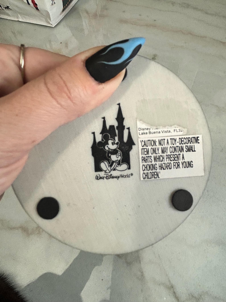 May include: A round, off-white Disney coaster featuring a black silhouette of a castle and Mickey Mouse. The coaster has a warning label and two black rubber feet. The text on the coaster reads "Walt Disney World".